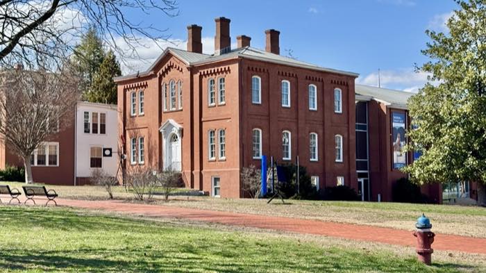 A red brick building with tall arched windows and a white doorway, a modern glazed wing to the right carrying an exhibition banner, lawn and brick path in front.