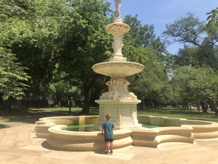 A tiered carved marble fountain with a statue on top playing into a scalloped basin, a small child watching from the rim, mown park lawn and mature trees behind.