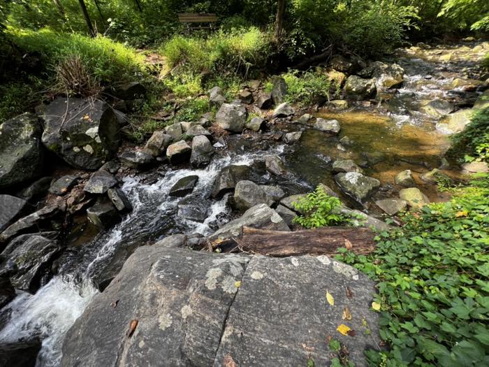 A rocky stream dropping over a low ledge into a clear pool, moss-green boulders on both banks and a wooden bench up on the far side.