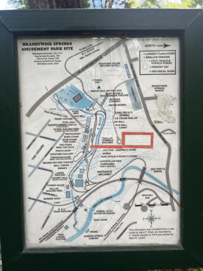 A framed New Castle County interpretive sign showing a labeled map of the 1911 Brandywine Springs amusement park, with the roller coaster, carousel, dance hall on the lake, trolley line and dozens of other attractions drawn over the present-day roads and creeks.