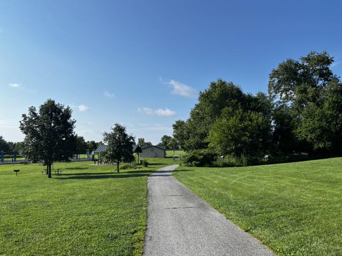 An open mown field with a paved path curving through it, picnic pavilions and a small building to the left, and play equipment in the distance.