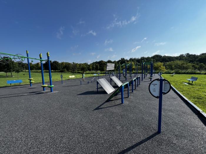 A long outdoor ninja obstacle course on black rubber surfacing: balance beams, angled boards, hanging rings and climbing frames, with two children on it and open playing fields either side.
