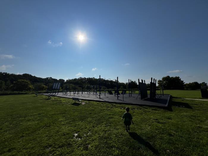 A wide view across mown grass to a long ninja obstacle course silhouetted against a bright sky, with a small child walking towards it.