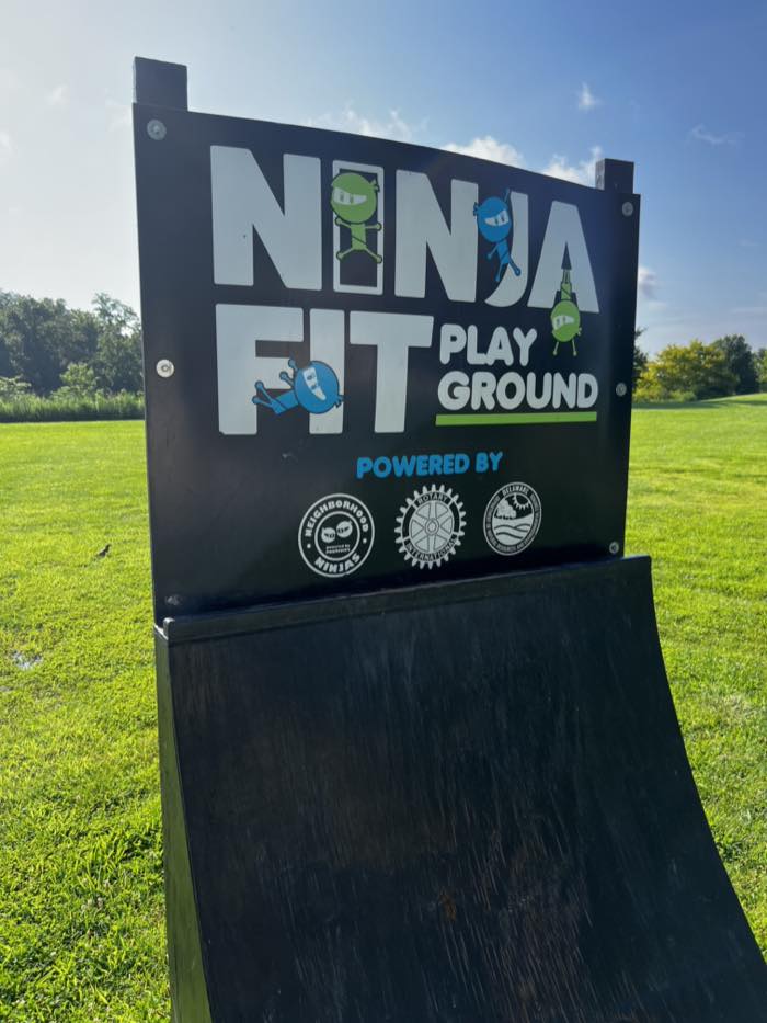 A sign reading NINJA FIT PLAY GROUND in white on black, with cartoon ninja characters, standing at the end of a curved black warped wall on mown grass.