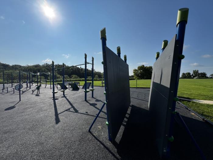 Two tall angled climbing boards with green hand-holds in the foreground of a ninja obstacle course, with monkey bars and balance equipment behind and a child in the middle distance.