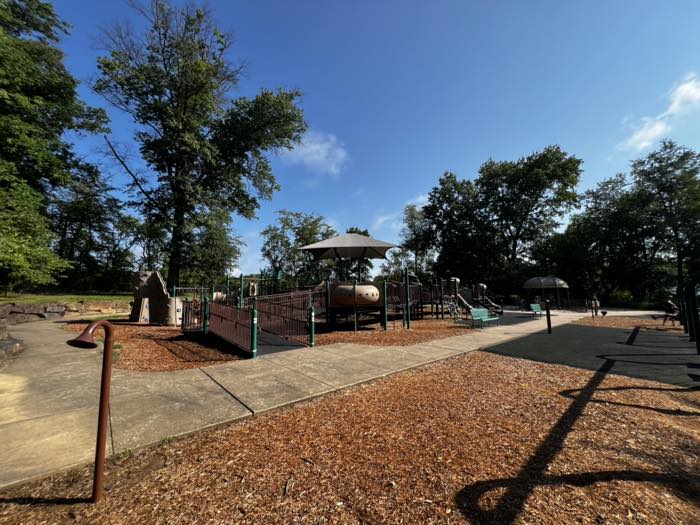 A wide view of a large playground with brown ramps and railings up to elevated decks, a shade canopy over the center, wood-chip surfacing and concrete paths.