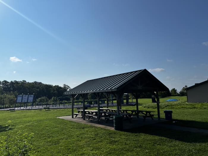 A green-roofed timber picnic pavilion with picnic tables under it, on grass beside a paved path, with the ninja course visible in the distance behind.