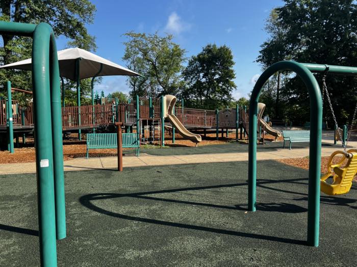 Playground swings in the foreground on dark poured-rubber surfacing, with a long ramped play structure, slides, shade sails and benches behind.