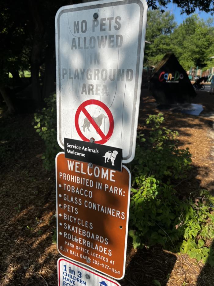 Signs on a post reading No Pets Allowed In Playground Area with a crossed-out dog, a plaque reading Service Animals Welcome, and a brown sign listing what is prohibited in the park.