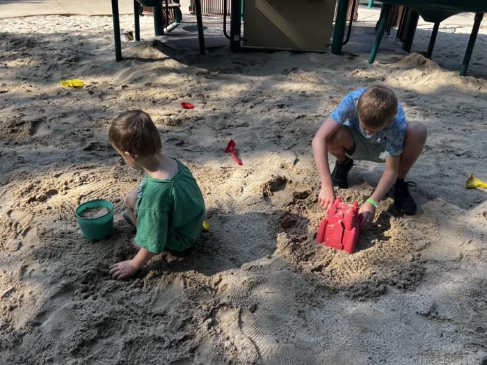 Two children crouched in the playground's sand area, seen from behind and above, one filling a bucket and one turning out a red sandcastle mould, with plastic spades scattered around.