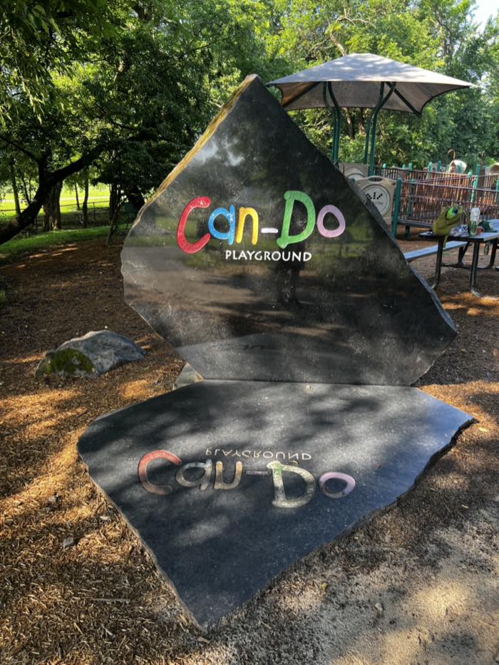 A low stone slab set in mulch, carved and painted with the words Can-Do Playground in red, blue, yellow, green and pink letters, with play equipment behind it.