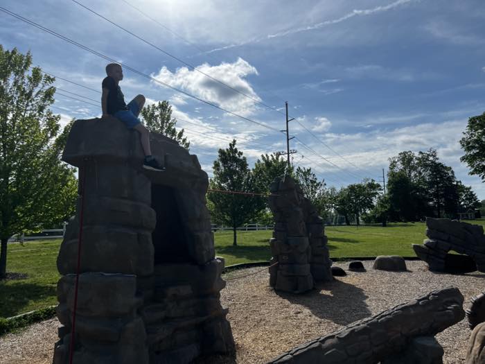 A child sitting on top of a cast-concrete rock formation with a cave beneath it, part of a climbing playground on wood chips, with more rock pinnacles and a fenced pasture behind.