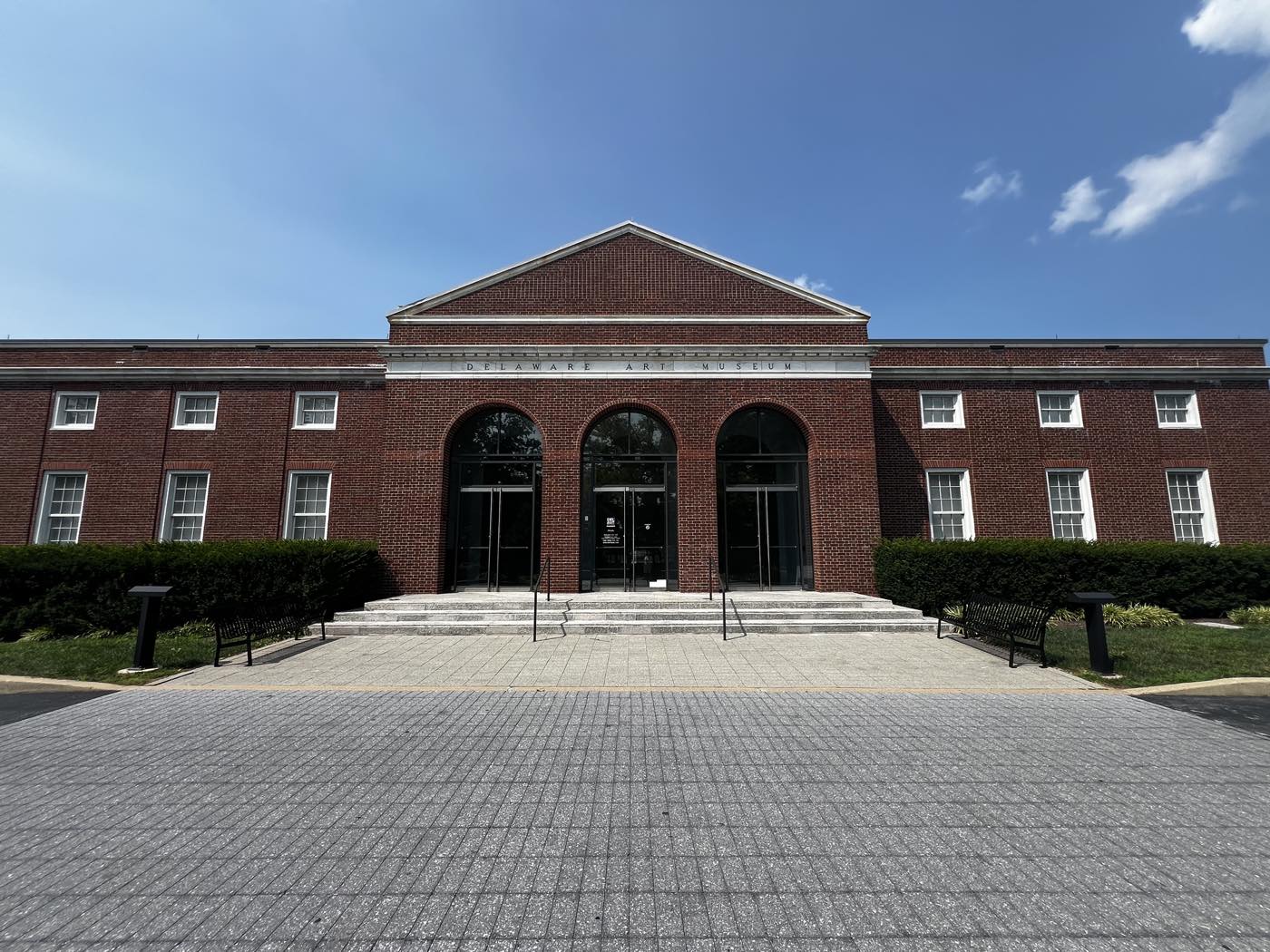 The Delaware Art Museum's red brick facade with three tall arched glass entrances under a pediment, DELAWARE ART MUSEUM carved in the stone band above them, benches either side of a wide paved forecourt.