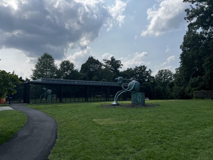 A long low glass-walled pavilion beside mown lawn, with a large green-patinated bronze sculpture of a stylised figure on a cylindrical plinth in the foreground and a tarmac path curving past.