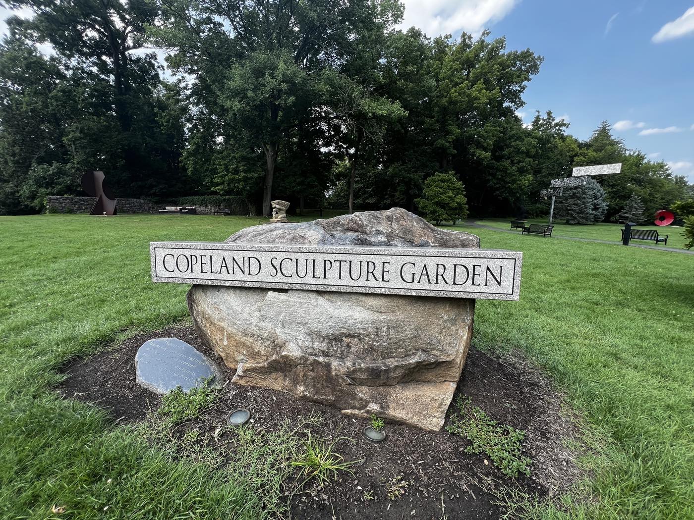 A granite sign reading COPELAND SCULPTURE GARDEN mounted on a boulder, with mown lawn, benches and several large sculptures among trees behind it.