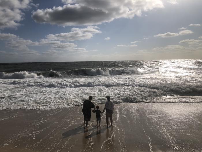 Three figures in silhouette standing at the edge of the surf with their backs to the camera, facing a line of breaking waves under a wide cloudy sky, sun glare on the water to the right.