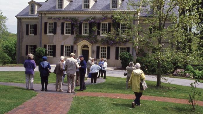 A large stone house with black shutters and wisteria over the front, a tour group of about a dozen people standing on the lawn and brick path in front of it.