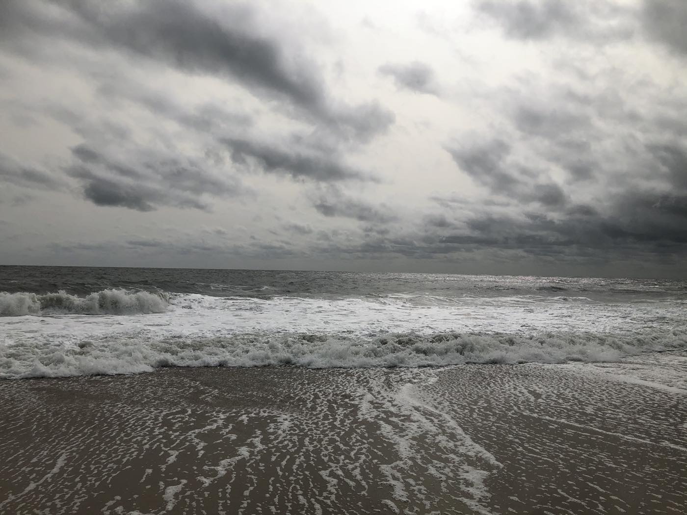Surf breaking on an empty brown-sand beach under a heavy gray overcast, with a long low line of white water running the width of the frame.