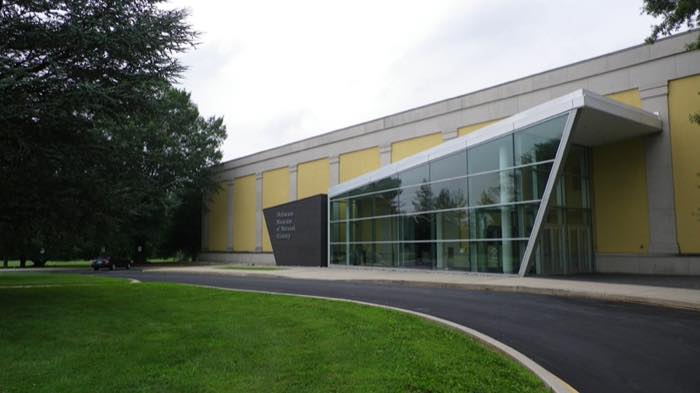 A long low museum building with yellow panels and a sharply angled glass entrance canopy, lawn and driveway in front.