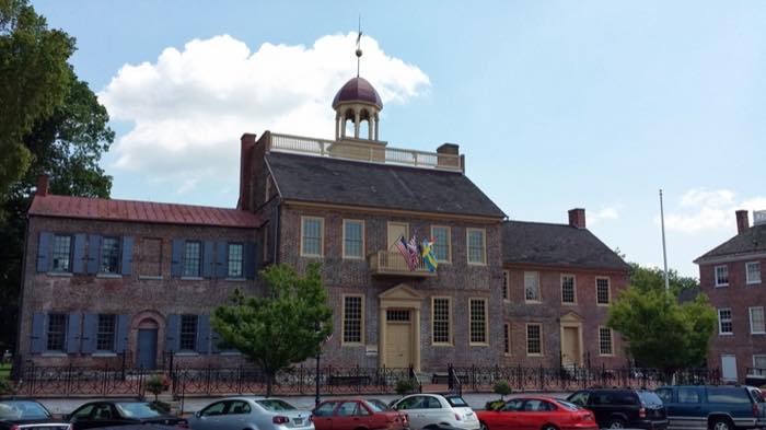 A brick colonial courthouse with a white cupola and balcony, three flags flying from it, cars parked along the street in front.