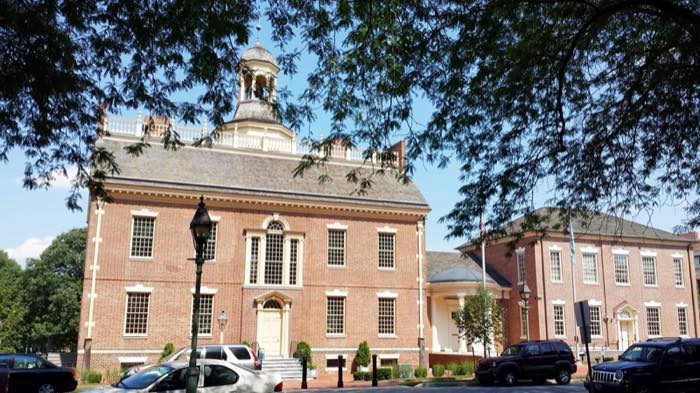 A red brick Georgian state house with a white cupola and arched window, framed by a tree, on Dover's green.