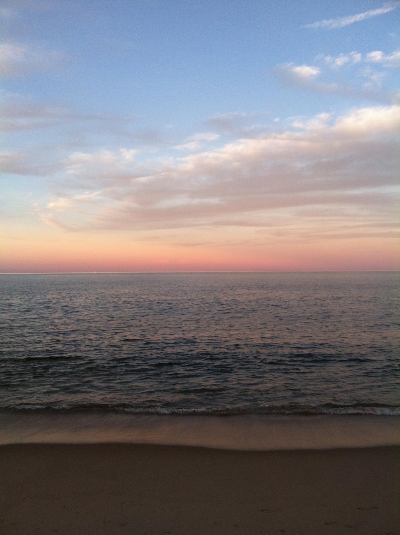 A calm sea at first light under a pink and pale-blue sky, with a thin line of surf and empty wet sand across the bottom of the frame.
