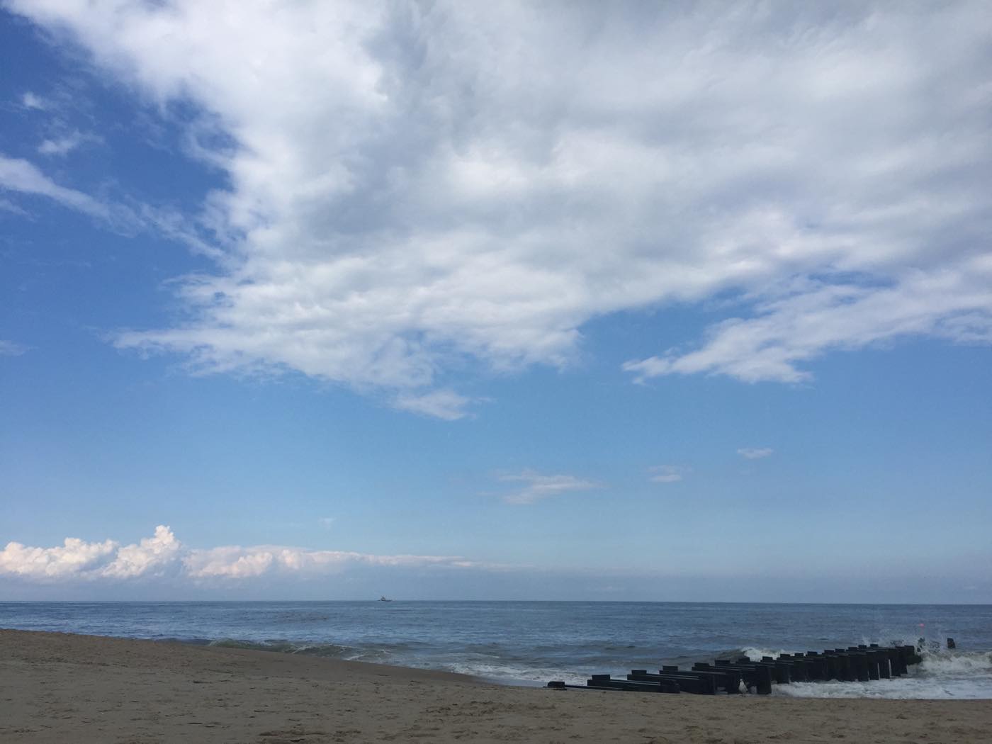 A wide empty sand beach under a tall blue sky with high white cloud, a black timber groin running out into the sea at the right and a small boat on the horizon.