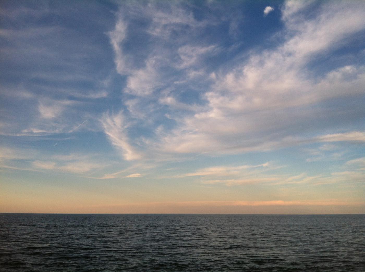 An open sea horizon at the end of the day under streaked cirrus cloud, the sky shading from blue to warm cream just above the waterline.
