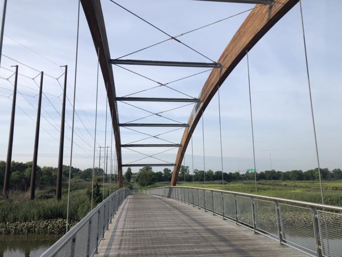 Looking along a wide timber-decked footbridge with a laminated timber arch overhead and steel cables, marsh and power lines on both sides.