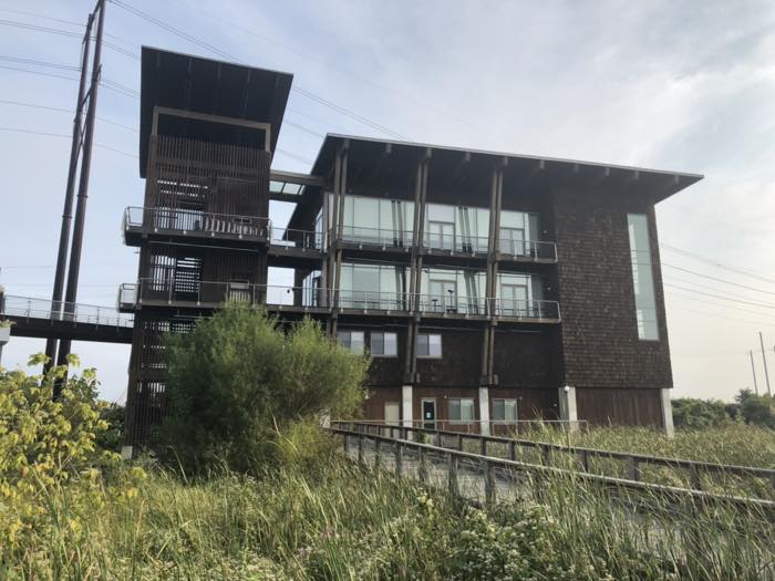 A four-story building clad in dark shingles and timber slats with a tall open stair tower beside it, seen from below across tall marsh grass.