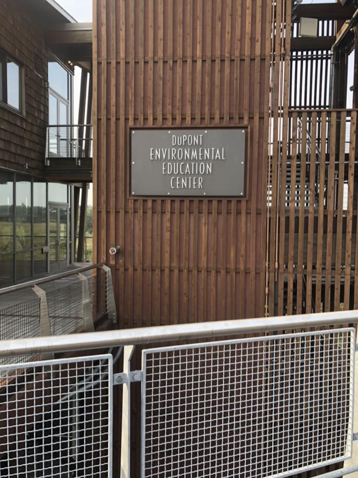 A gray sign reading DuPont Environmental Education Center mounted on a slatted timber wall, with steel mesh walkway rails in front.