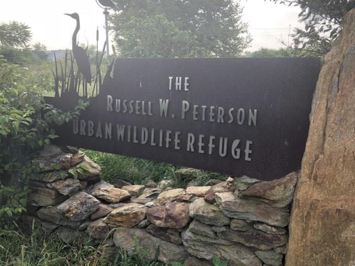 A dark metal sign on a dry stone wall reading The Russell W. Peterson Urban Wildlife Refuge, with a cut-out heron and cattails at its left end.