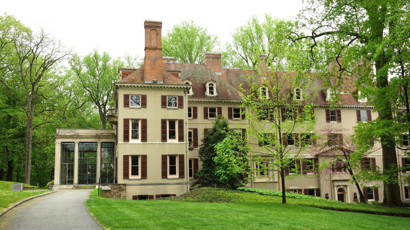 A very large cream-coloured country house with brick chimneys and a glazed conservatory wing, surrounded by mature trees and lawn in spring.