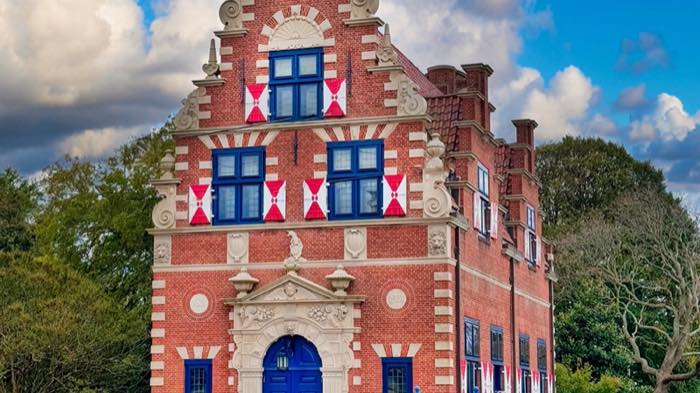 An ornate Dutch-style brick building with a stepped and scrolled gable, red and white shutters, and a blue front door under carved stonework.
